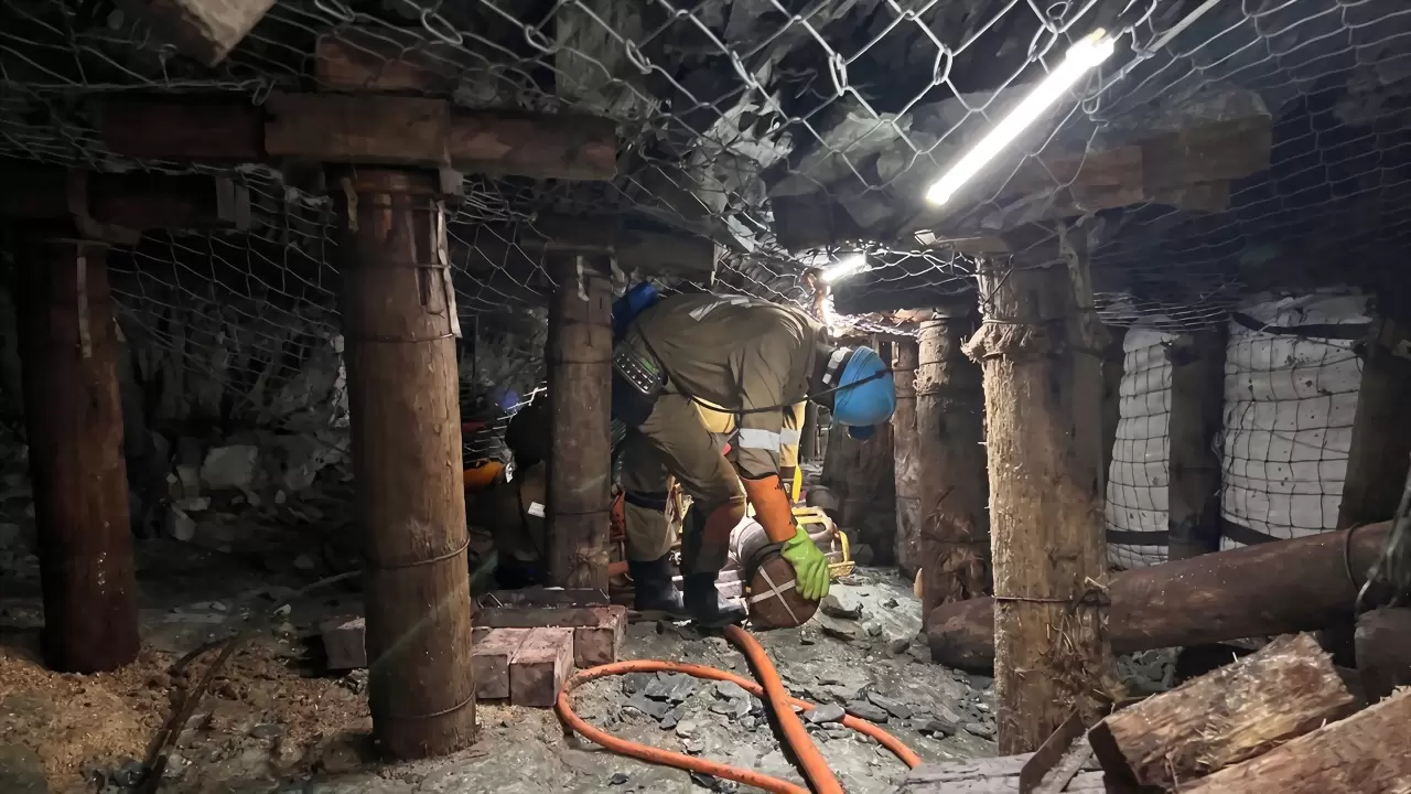 A miner in overalls and a hard hat crouching beneath timber mine supports, with a bright white LED strip running along the roof above